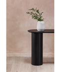 Black cylindrical table with a white vase and green plant against a beige wall.
