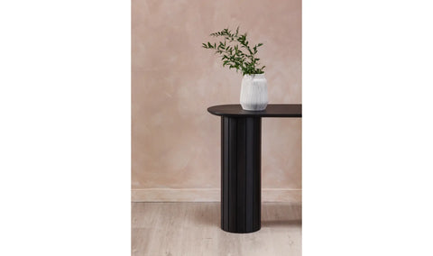 Black cylindrical table with a white vase and green plant against a beige wall.