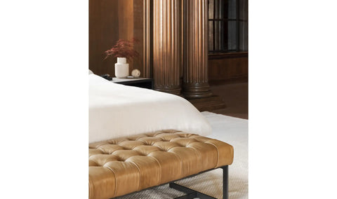 Brown tufted bench in front of a bed with white bedding in a bedroom setting.